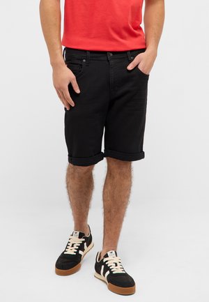 Mann trägt schwarze hochgekrempelte Shorts, rotes T-Shirt und schwarze Turnschuhe mit weißen Schnürsenkeln und Gummisohlen, steht mit einer Hand in der Tasche.