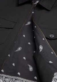 Veste noire avec une fermeture éclair partiellement ouverte qui dévoile une doublure en tissu noir décorée de motifs cachemire blancs.