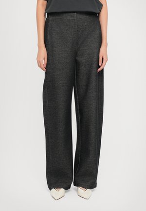 Femme portant un pantalon large gris foncé et des chaussures blanches à bout pointu, debout devant un fond uni.