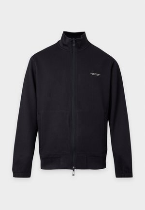 Homme debout, face à l'objectif, portant une veste noire zippée, un jean bleu clair déchiré et des baskets blanches sur un fond blanc uni.