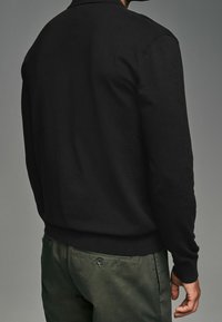 Zwarte gebreide trui met een ronde halslijn, lange mouwen en geribde manchetten, gecombineerd met olijfgroene broek. Zachte textuur, minimalistisch ontwerp.