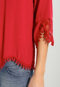 Blusa rossa con dettagli in pizzo e bordatura a pompon su maniche e orlo. Tessuto leggero, caratterizzata da una vestibilità rilassata e uno stile informale.