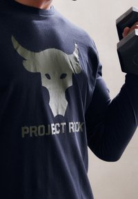 Person, die ein dunkles Shirt mit einem weißen Bullenkopf und dem Text "PROJECT ROCK" trägt, hält in einer Trainingsumgebung eine schwarze Hantel.