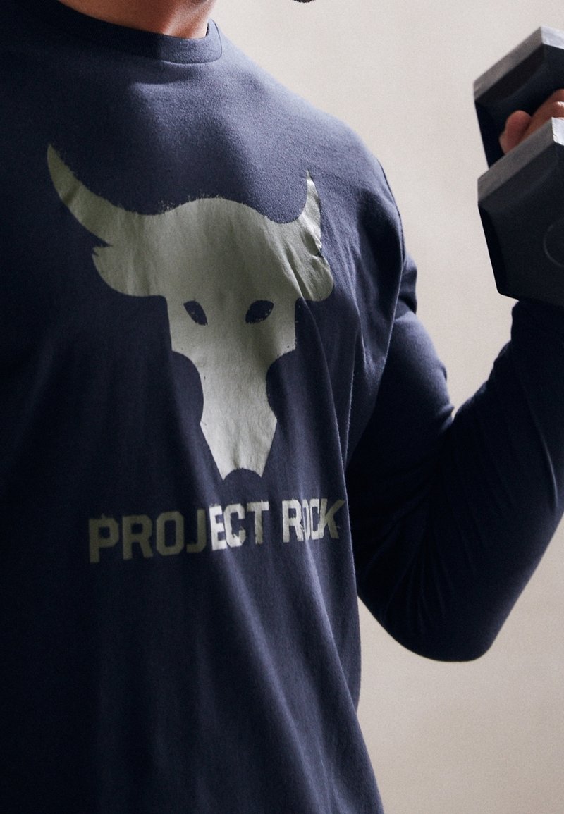 Osoba oblečená v tmavém triku s bílou lebkou býka a textem "PROJECT ROCK", držící černou činku v posilovacím prostředí.