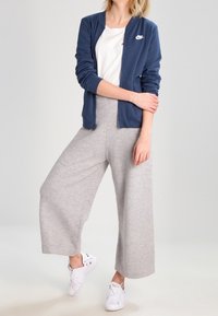 Kvinna i marinblå zip-up Nike-jacka, vit t-shirt, grå vida byxor och vita sneakers, stående med ett ben något upplyft.