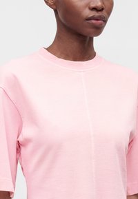 Personne portant une chemise rose clair à manches courtes avec un col rond et une couture verticale centrale sur un fond uni.
