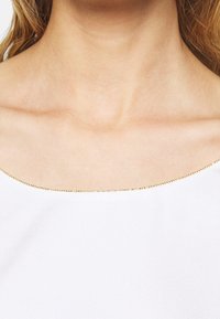 Haut blanc avec un décolleté arrondi et une bordure en perles dorées. Le tissu a une texture lisse, adapté pour les occasions décontractées ou formelles.