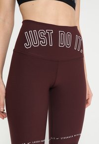 Leggings bordeaux a vita alta con testo bianco "JUST DO IT." sulla cintura. Presentano un materiale liscio e elastico e cuciture piatte.