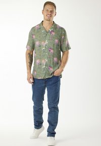 Camicia verde a maniche corte con motivo colorato di palme, con bottoni frontali, abbinata a jeans blu e sneakers bianche.