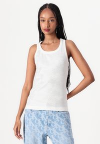 Wit tanktop met een netpatroon, met een ronde halslijn en brede banden. Gecombineerd met lichtblauwe, gemusterde jeans.