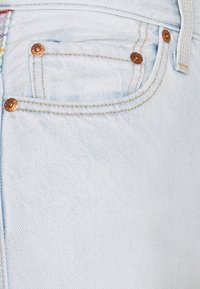 Gros plan sur un jean en denim bleu clair montrant la poche avant, les passants de ceinture et les rivets en cuivre avec des détails de couture jaune.