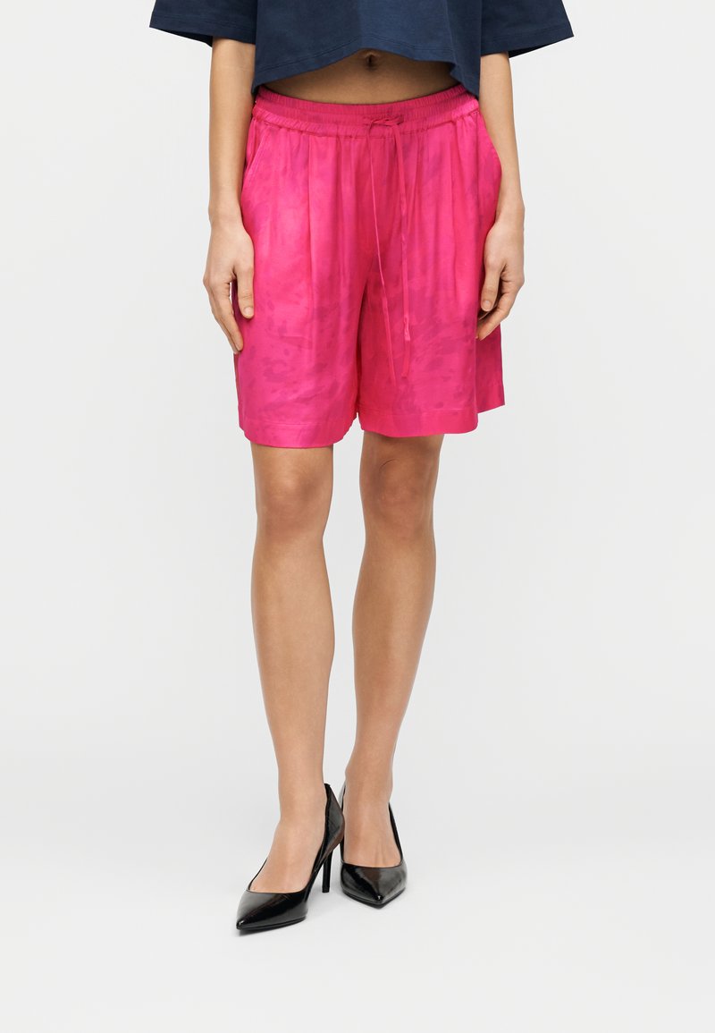 Ροζ σατέν σορτς με ελαστική μέση και κορδόνι. Μήκος μέχρι το γόνατο με ομαλή υφή και διακριτικό σχέδιο. Συνδυασμένο με μαύρα τακούνια.