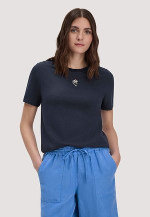 Frau mit braunem, welligem Haar trägt ein marineblaues T-Shirt mit kleiner weißer Blumenstickerei und locker sitzende hellblaue Hose mit Kordelzug, Hände in den Taschen.