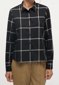 Chemise à carreaux noir et beige à boutons, à manches longues, avec des boutons noirs et un col classique, portée sur un pantalon beige.