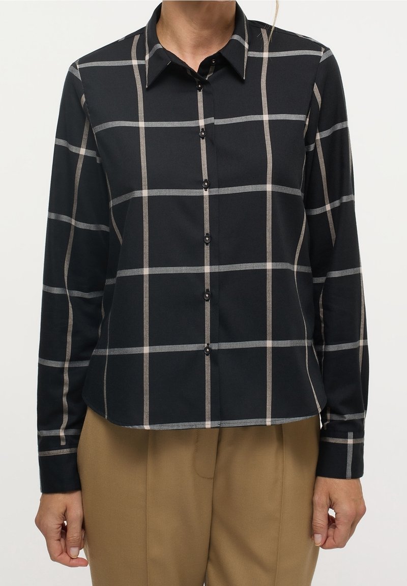 Chemise à carreaux noir et beige à boutons, à manches longues, avec des boutons noirs et un col classique, portée sur un pantalon beige.