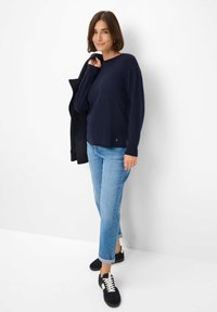 Vrouw die tegen een witte muur staat, gekleed in een marineblauwe trui, lichtblauwe jeans, zwarte sneakers en een zwarte jas over haar schouder houdt.
