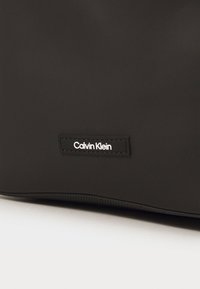 Svart Calvin Klein-veske laget av glatt materiale, med rektangulær fasong og en merket, sydd etikett. Enkel og minimalistisk design.