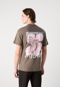 Brown cotton t-shirt featuring a pink abstract graphic with the text "DIGITAL MYSTERY" in white, short sleeves, and a relaxed fit.