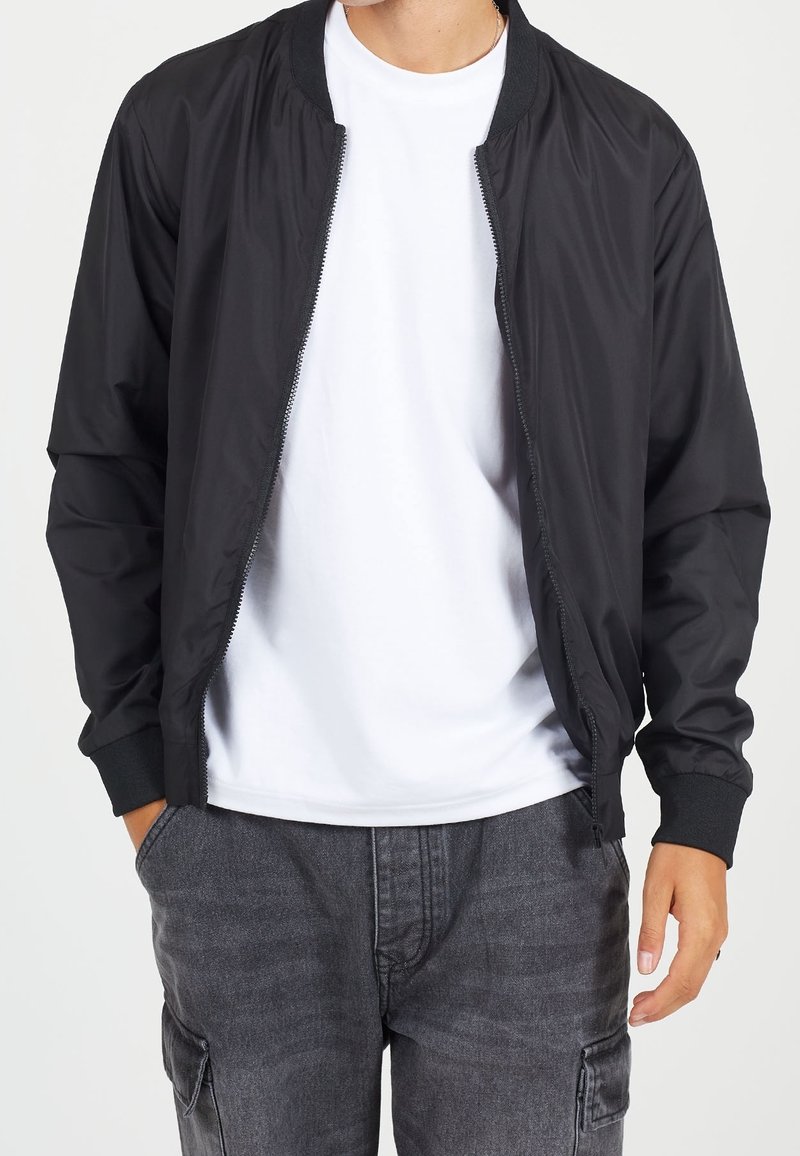 Black bomber jacket made of smooth fabric with a front zipper, ribbed cuffs, and a round neckline, worn over a white t-shirt.