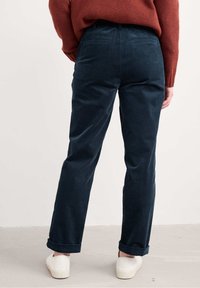 Jūras zila korduroja bikses ar taisnām kājām un uzvelkamām malām, ar aizmugurējām kabatām un gludu tekstūru. Vilktas kopā ar baltām slip-on kurpēm.