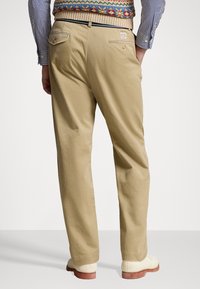 Bruna bomullstrousers med avslappnad passform, med bakfickor och en logotypetikett. Kommer med krämfärgade skor och en mönstrad stickad tröja.