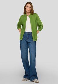 Grüne, gerippte Bomberjacke mit rundem Ausschnitt, getragen über einem weißen Hemd; kombiniert mit hoch taillierten, blauen Jeans mit weitem Bein und silbernen Schuhen.