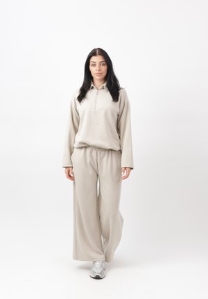 Frau mit langen dunklen Haaren, die ein beiges, locker sitzendes Oberteil und weite Hosen trägt, geht vorwärts vor weißem Hintergrund mit silbernen Turnschuhen.