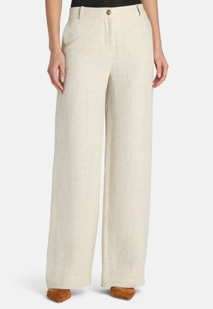 Moteris dėvi aukšto liemens, plačias smėlio spalvos lino kelnes su priekiu sagomis ir diržo kilpomis, derina su rudais, smailiais batais.