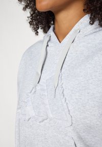 Lichtgrijze hoodie met een gestructureerde afwerking, voorzien van een witte gerimpelde design op de voorzak en grijze koordjes bij de hals.