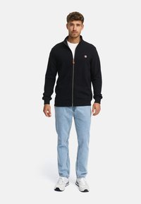 Hombre de pie de frente, vestido con chaqueta negra con cremallera, camiseta blanca, jeans de mezclilla azul claro y zapatillas blancas, sobre un fondo liso claro.