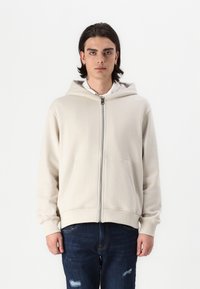 Beige zip-up hooded sweatshirt with a front pocket, cotton blend fabric, and ribbed cuffs. Worn over a white collared shirt.