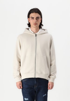 Kremo spalvos zip-up su kapisonu megztinis su priekinėmis kišenėmis, pagamintas iš medvilnės mišinio, su rausvais rankogaliais. Dėvimas virš baltos marškinėlių su apykakle.