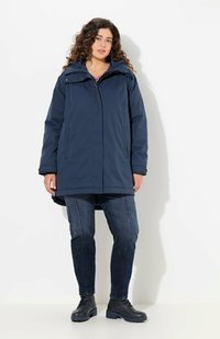 Giacca impermeabile blu navy con cappuccio, zip frontale e tasche con patta. Abbinata a jeans in denim scuro e stivali neri con lacci.