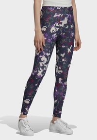 Kvinna i högmidjade, mörkblommiga leggings och vita sneakers, stående mot en enkel ljus bakgrund.