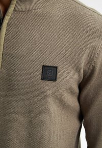 Pulover tricotat dintr-un material brun texturat, având un guler cu fermoar pe jumătate și un patch cu logo negru pe piept.