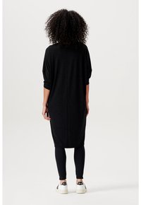 Vestido negro de gran tamaño con mangas de tres cuartos, dobladillo redondeado y una textura suave, combinado con leggings negros y zapatillas blancas.