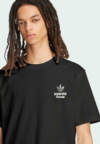Black t-shirt featuring a small white Adidas logo on the left chest, with the text "superstar PARIS" below the logo. Short sleeves, smooth fabric.