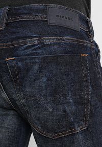 Tmavě modré džíny z denimu s texturovaným povrchem, viditelným šitím v oranžové barvě, přizpůsobenými zadními kapsami a černým koženým logem.