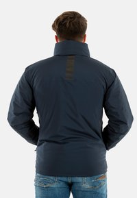 Veste isolante bleu marine avec un col haut, un logo à l'arrière, et une texture lisse, associée à un jean en denim bleu.