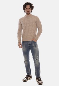 Uomo con capelli ricci che indossa un maglione beige a trecce, jeans blu strappati e sneakers marroni e bianche, in piedi con una mano in tasca.