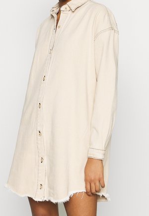 Osoba ubrana w dużą beżową sukienkę-koszulę zapinaną na guziki, z postrzępionym dołem i długimi rękawami, stojąca na tle jednolitego jasnego tła.