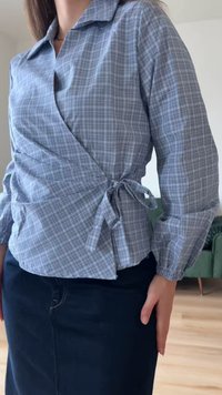 Γυναίκα που φοράει μπλε καρό μπλούζα τύπου wrap με φιόγκο δεμένο στο πλάι και σκουρόχρωμη φούστα, στέκεται σε εσωτερικό χώρο με μια πράσινη καρέκλα στο φόντο.