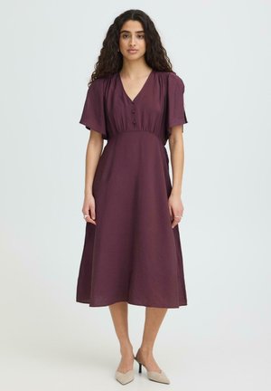 Donna con lunghi capelli ricci che indossa un vestito viola scuro corto a maniche corte fino al ginocchio e scarpe beige con tacchi alti, in piedi davanti a uno sfondo semplice.