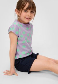 Lila t-shirt met groene palmbladprint, korte mouwen; gecombineerd met donkerblauwe shorts. Kind dat op de vloer zit, glimlachend.