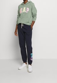 Sudadera verde con capucha y logo "GAP" en colores pastel; combinada con pantalones deportivos azul marino con un logo degradado y zapatillas blancas.