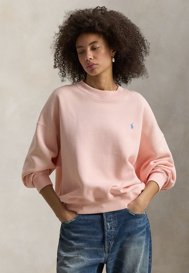 Ροζ φούτερ με στρογγυλή λαιμόκοψη και χαλαρούς ώμους. Διαθέτει ριμπ μανσέτες και τελείωμα, με μικρό μπλε λογότυπο στο αριστερό στήθος.