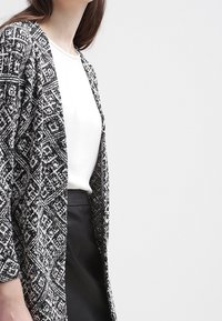 Czarny i biały kardigan w wzory z otwartym przodem, teksturowana tkanina, długie rękawy, nałożony na biały top i czarną spódnicę.