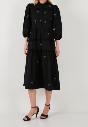 Robe noire mi-longue avec des manches bouffantes, fermée par des boutons sur le devant, au design à volants, ornée de broderies florales bleues et d'accents en fronces au niveau des coutures.