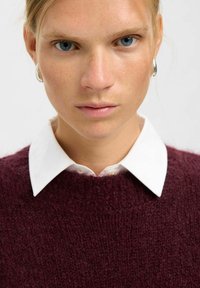 Burgunderfarbener Strickpullover mit strukturierter Oberfläche, kombiniert mit einem weißen Hemd mit Kragen darunter. Nahaufnahme der Gesichtszüge.