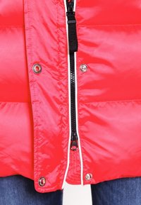 Gilet matelassé rouge avec fermeture éclair à l'avant, passepoil blanc contrastant et boutons-pression argentés. Présente une texture lisse et brillante.